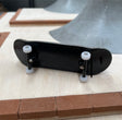 A black miniature fingerboard with two white wheels and metal trucks, placed on a wooden surface.