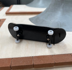 A black miniature fingerboard with two white wheels and metal trucks, placed on a wooden surface.